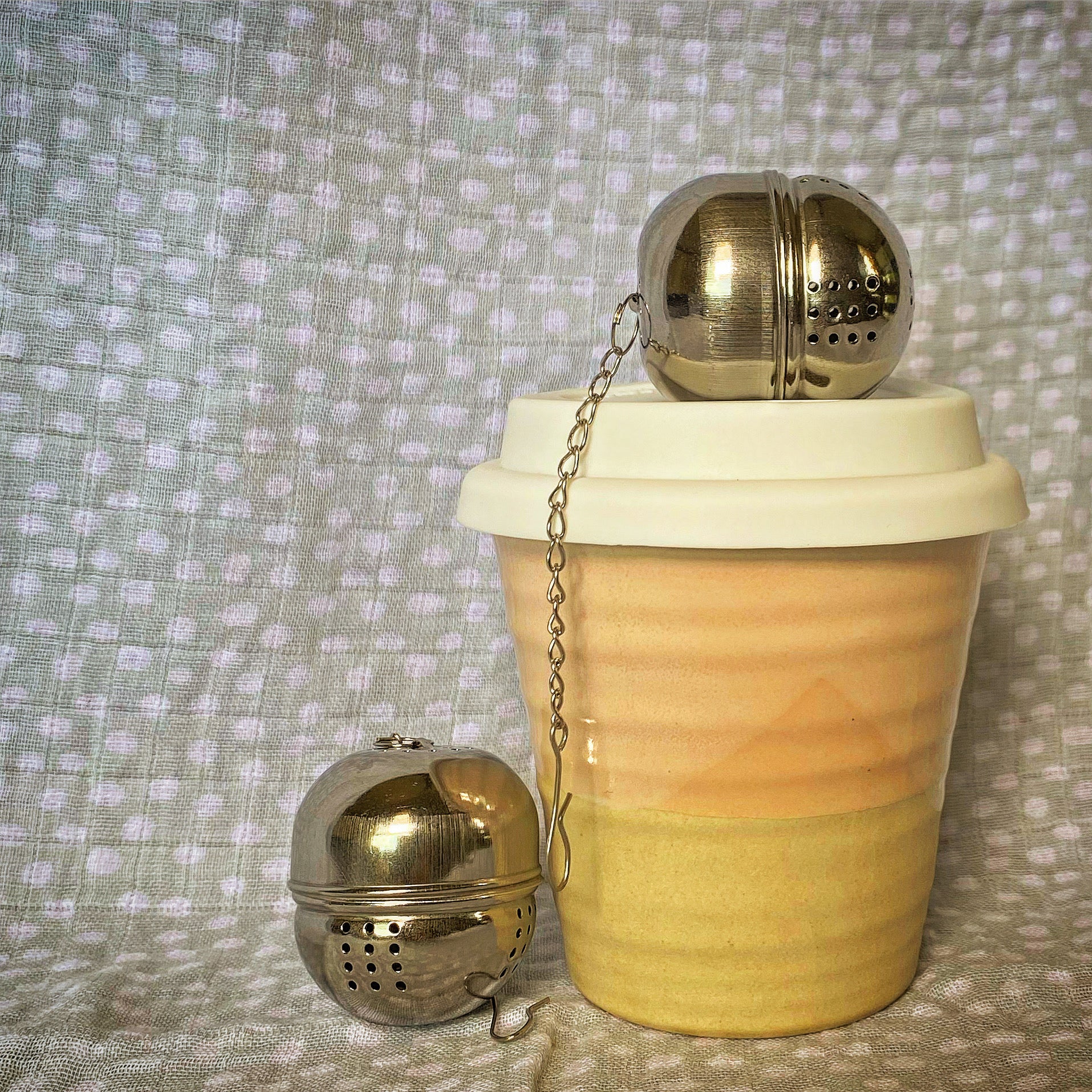 1 ceramic coffee keep cup with a stainless steel tea infuser ball on top and beside it. a green and white spotty blanket in the background